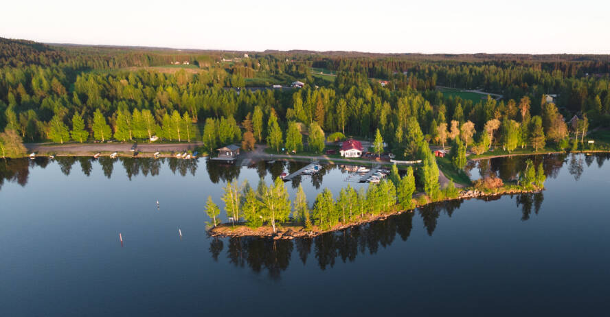 Järven rannalla sijaitseva satamapaviljonki kesäisessä maisemassa, kuvattuna ilmasta. Etualalla vehreä aallonmurtaja.