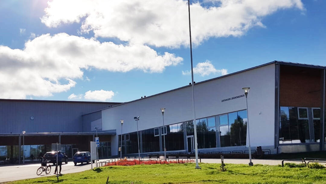 Aurikoinen kesä päivä. Pyöräilijä ja kaksi autoa liikuntahallin edessä.