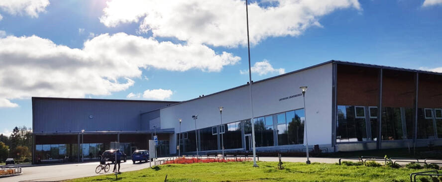 Aurikoinen kesä päivä. Pyöräilijä ja kaksi autoa liikuntahallin edessä.