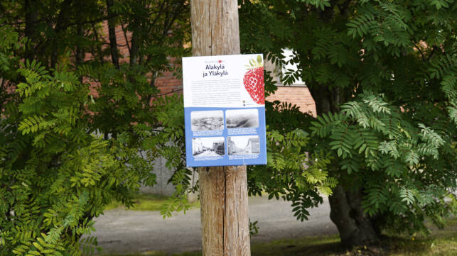 Kotiseutuopaste kiinnitettynä puutolppaan. Taustalla pihlajia.