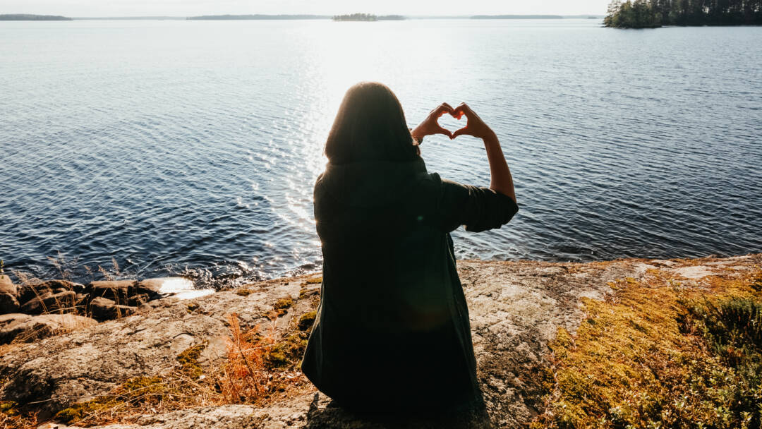 Rannalla vastavalossa istuu henkilö, joka muodostaa käsillään sydämen.