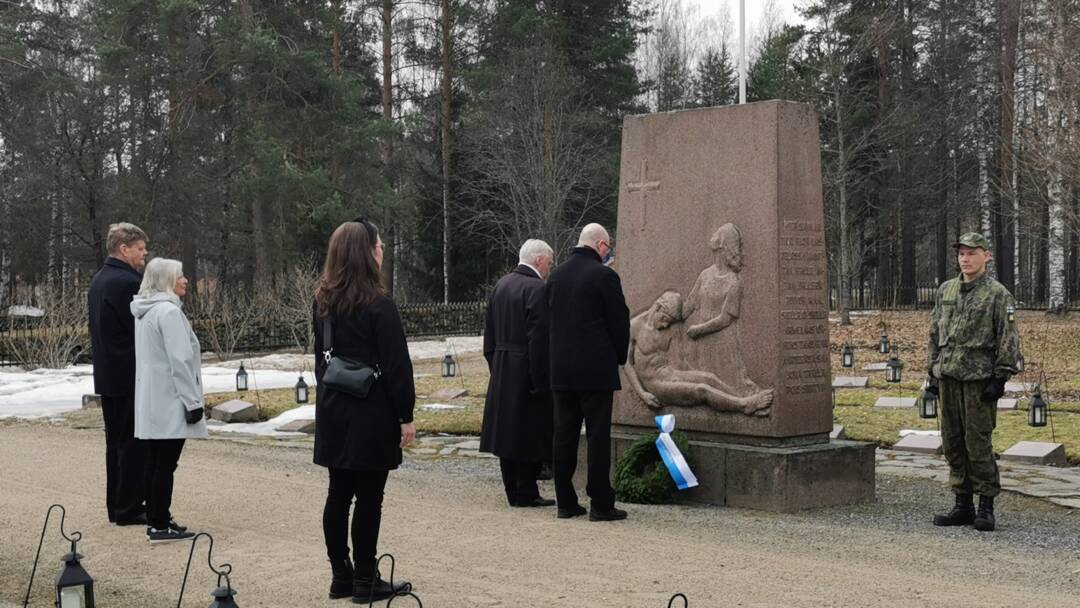 Kaksi henkilöä laskemassa seppelettä. Kaksi kunnioittamassa taka-alalla. Lisäksi kuvassa kunniavartio.