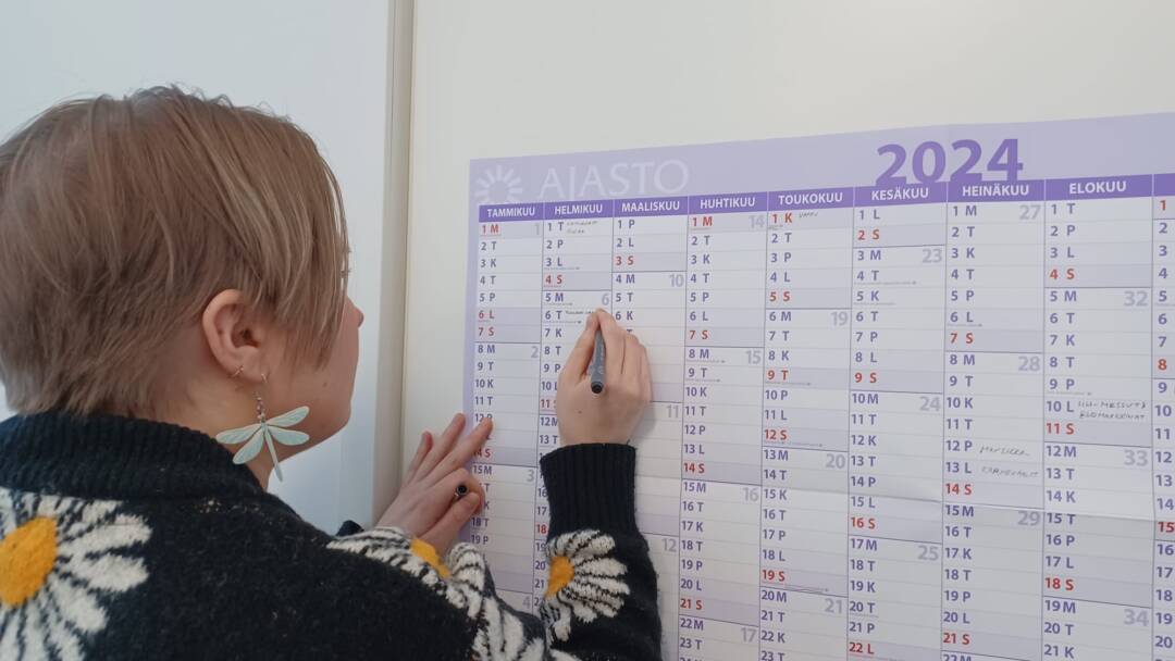 Nainen kirjoittaa seinällä olevaan kalenteriin