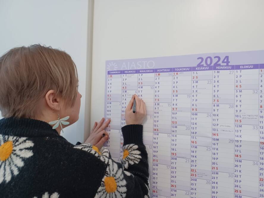 Nainen kirjoittaa seinällä olevaan kalenteriin