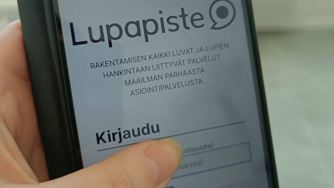 Kuvassa käsi, jossa älypuhelin. Älypuhelimen ruudussa Lupapisteen sivu.