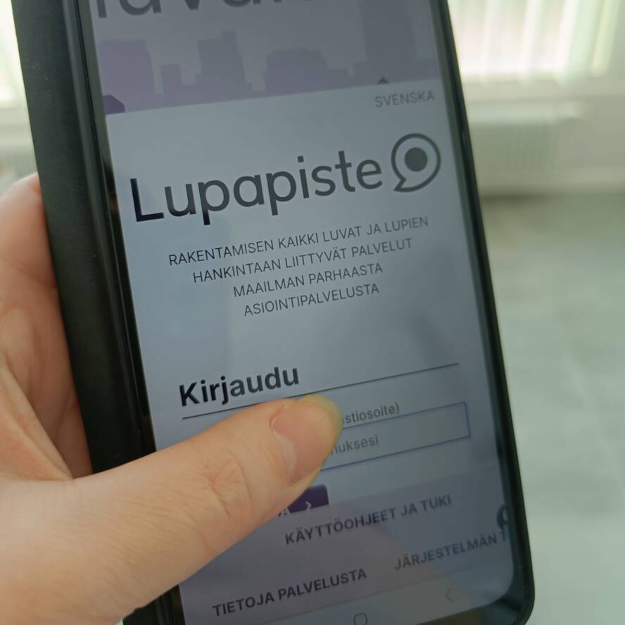 Kuvassa käsi, jossa älypuhelin. Älypuhelimen ruudussa Lupapisteen sivu.