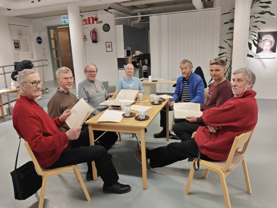 Joukko ihmisiä istuu pöydän ääressä ja katsoo kameraa.
