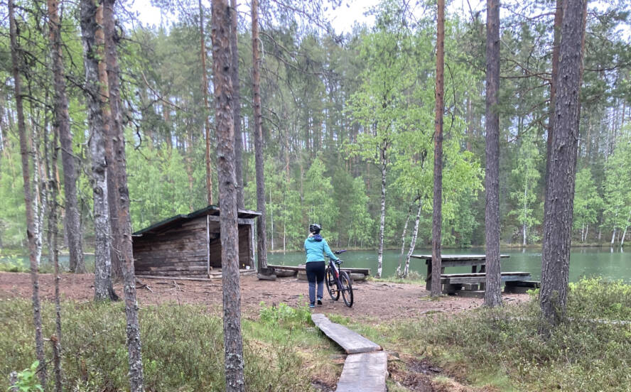 palolammen laavu, metsän keskellä, lammen rannalla, kuvan alalaidassa pitkospuut ja pitkospuiden päädyssä maastopyörää taluttava nainen sinisessä takissa.